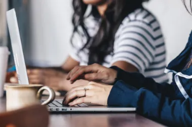 Two women sit at a table, each using a laptop, engaged in conversation and collaboration.